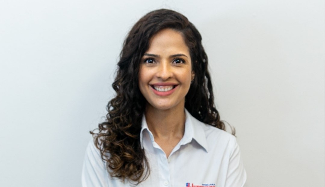 Uma mulher jovem está sentada à mesa de trabalho, sorrindo para a câmera. Ela tem cabelos lisos e escuros, na altura dos ombros, e usa óculos de armação escura. Ela veste uma camisa branca com o logotipo da 'Junqueira' no lado direito do peito e um crachá vermelho ao redor do pescoço. No fundo, há um telefone de mesa, um celular vermelho e um teclado de computador sobre a mesa. A expressão dela é amigável e profissional.